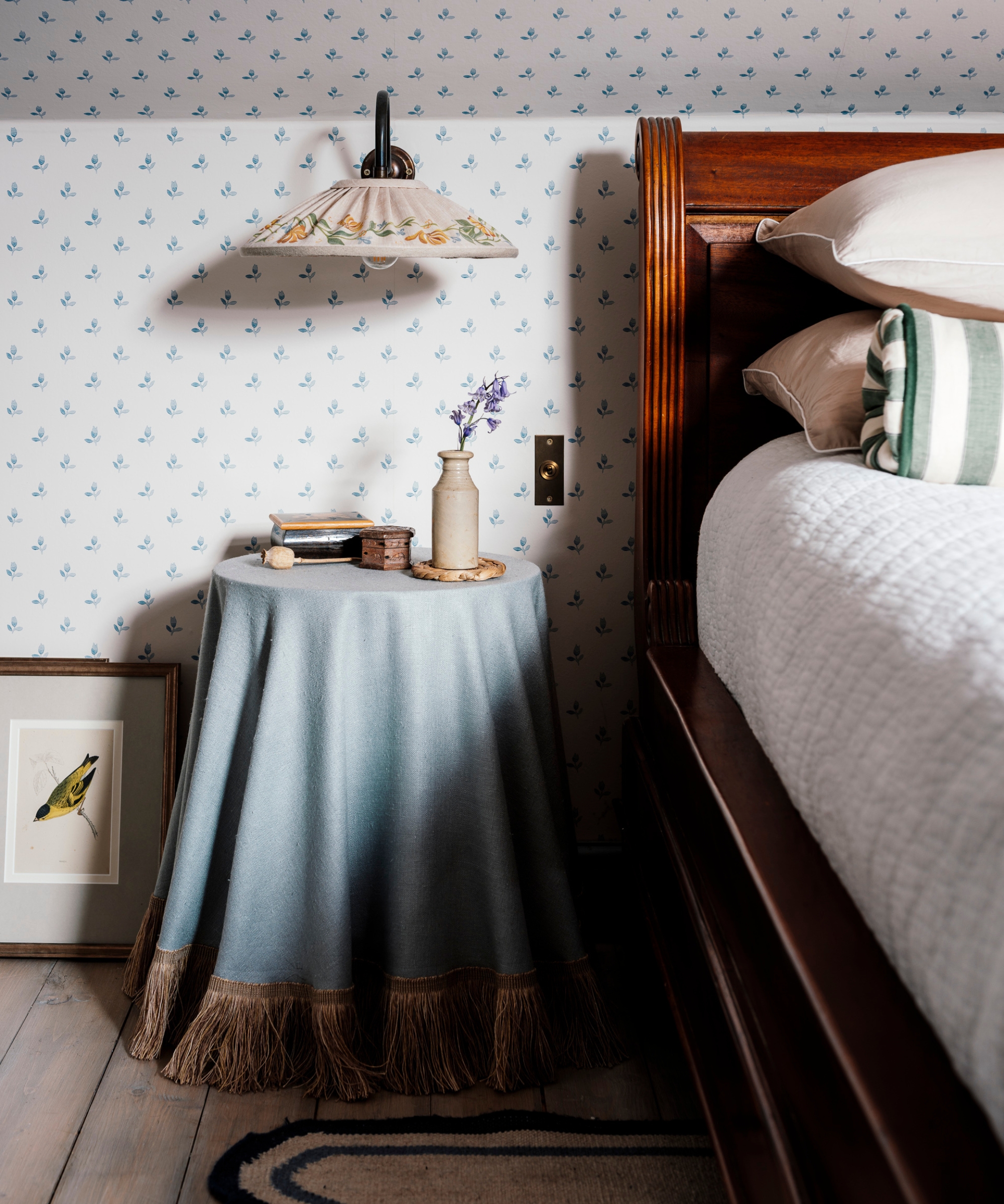 A floral embroidered wall lampshade above a blue fringed bedside table, beside a dark wooden bed and delicate blue botanical wallpaper.