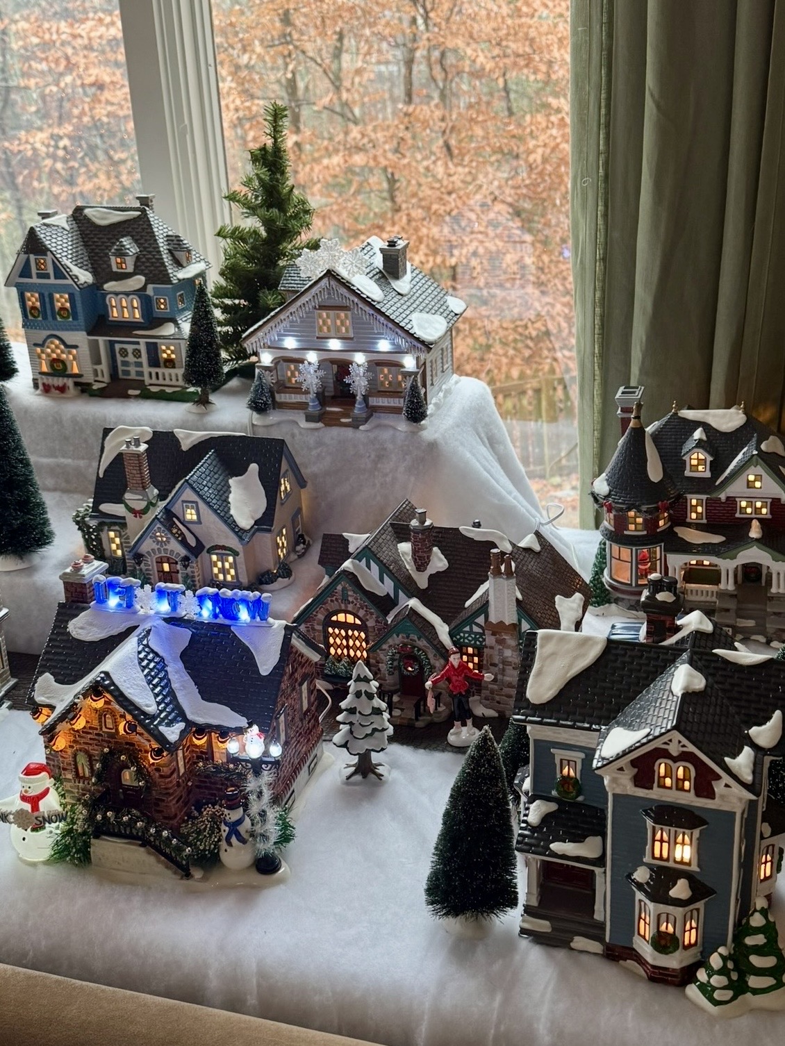 A miniature christmas village with ornate and lit up houses over multiple levels on surfaces of fake snow in front of a window with autumnal leaves on trees in the background