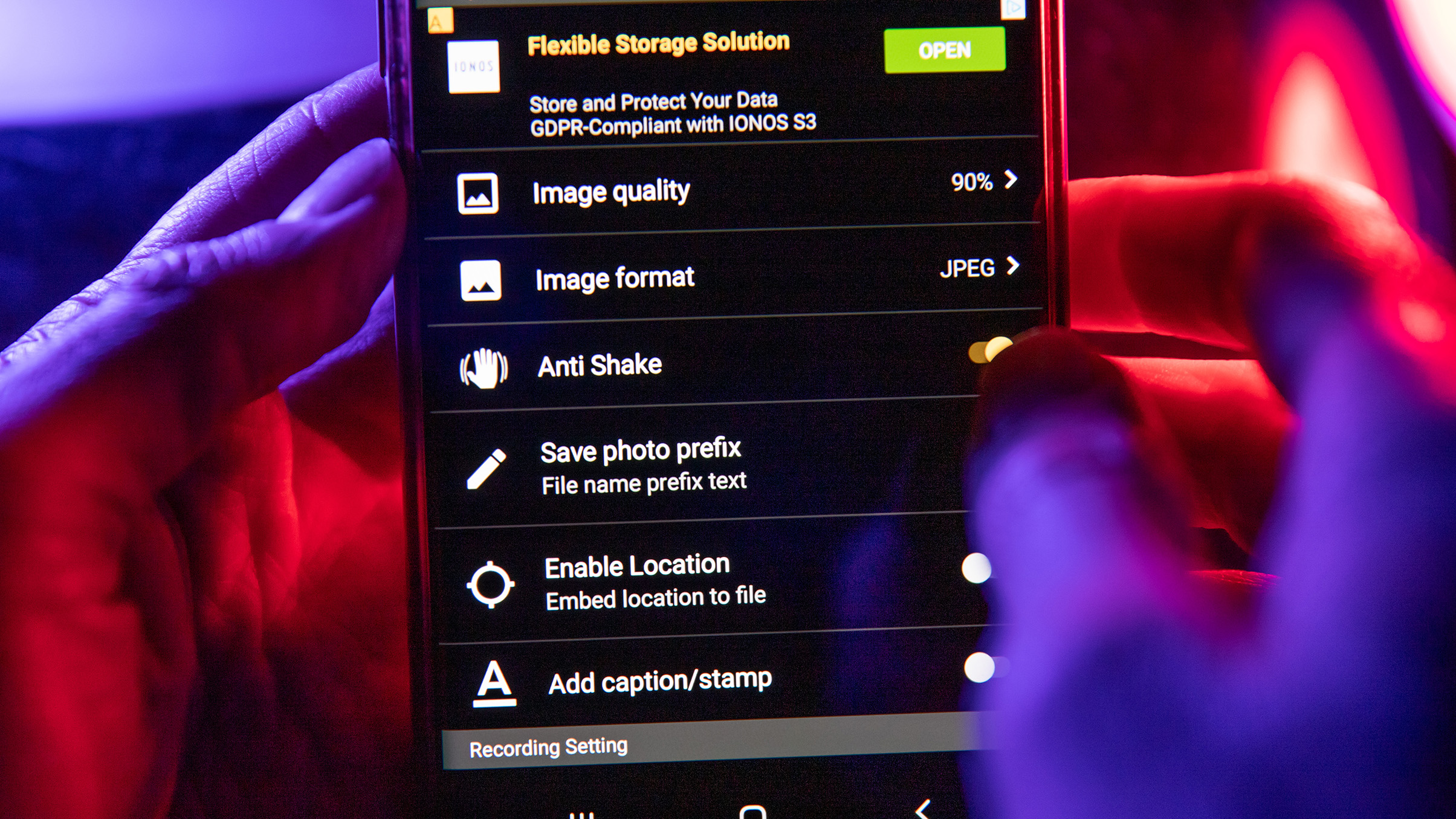 A close-up of a hand holding a smartphone displaying a settings menu for image capture options, illuminated with purple and red light