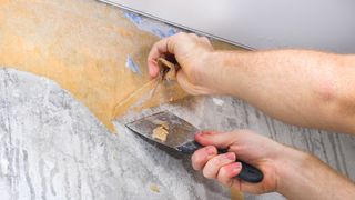 An image showing gray wallpaper being peeled off by hand using a putty knife