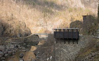 View of the black and timber service building which sits on a wall surrounded by rocks, a cliff and a creek