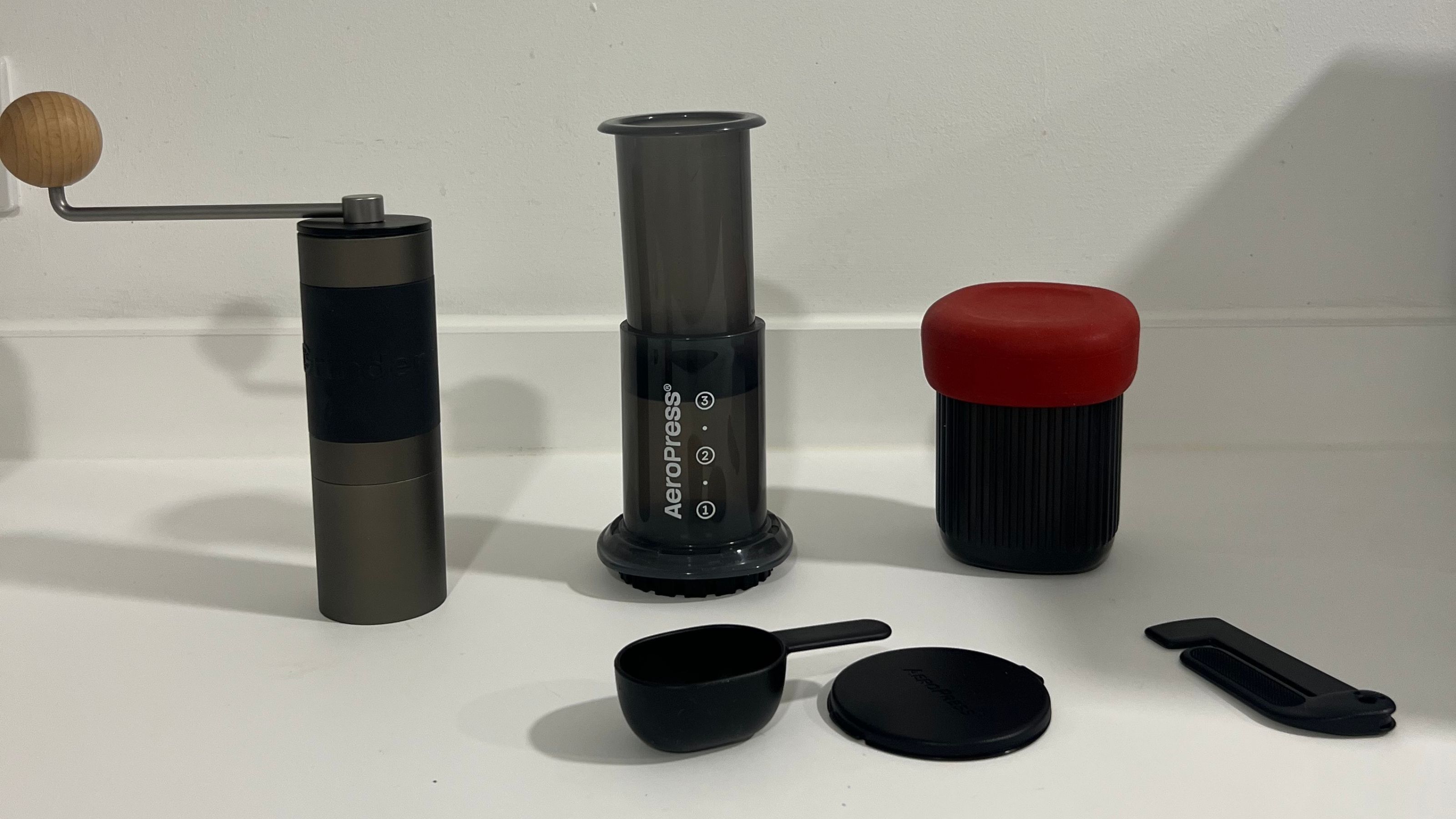 Image of a black AeroPress coffee machine and four of the AeroPress coffee making tools on a white countertop next to a black and gray manual coffee grinder.