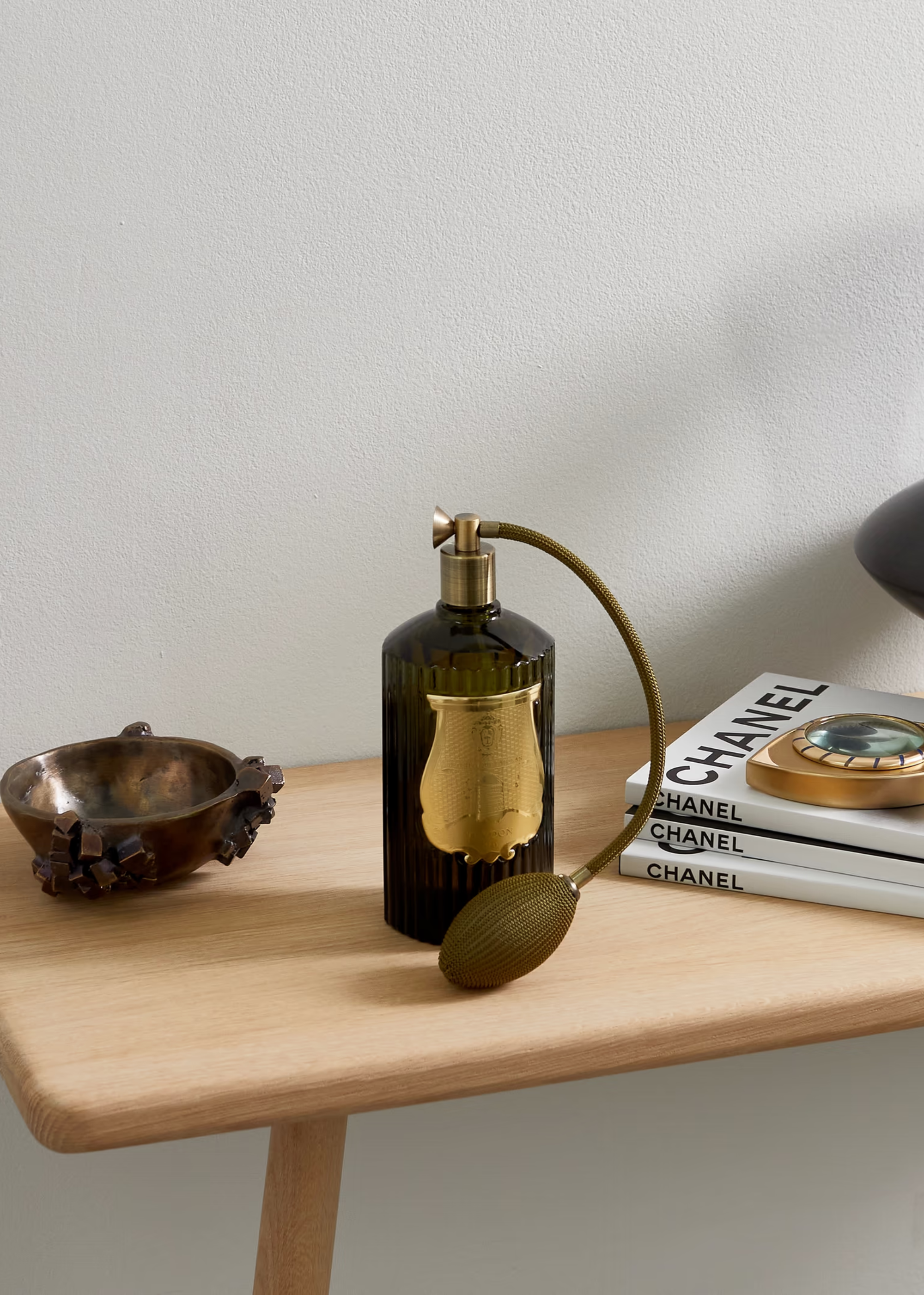 A bottle of Madurai Room Spray from Trudon on a wood side table beside a stack of Chanel magazines and a brass bowl