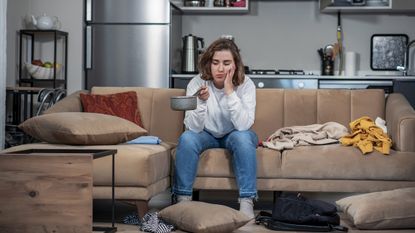 View of an adult female who is holding a pan. Because the ceiling is leaking water. Everything’s messed up. She is sitting on a sofa. She is in the kitchen. She is wearing a white- long-sleeved shirt and blue jeans. insurance concept.