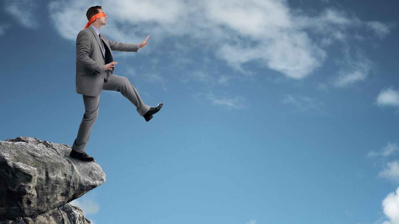 A blindfolded businessmen is about to step off the edge of a cliff.