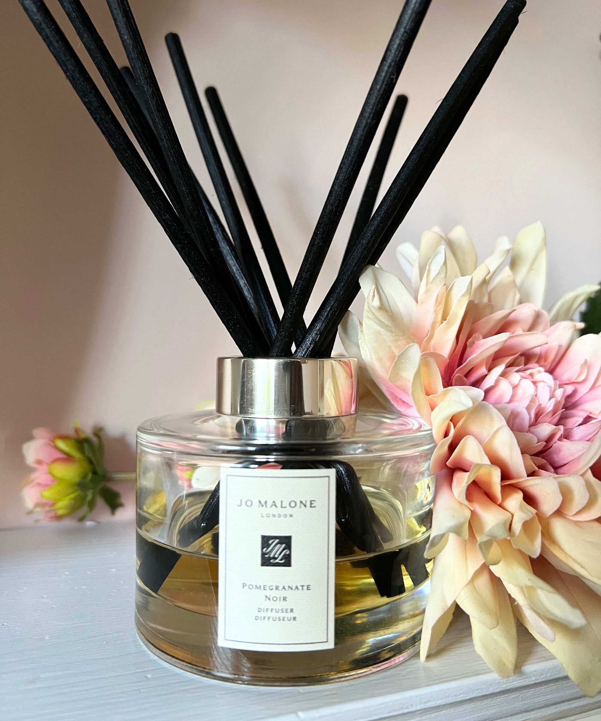 Jo Malone pomegranate noir diffuser stood on a white mantelpiece with pink flowers and pink wall behind