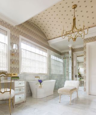 Large luxurious bathroom with cream patterned wallpapered ceiling and walls, white bath, glass shower, seat, marble flooring and wall tiles, gold chandlier,