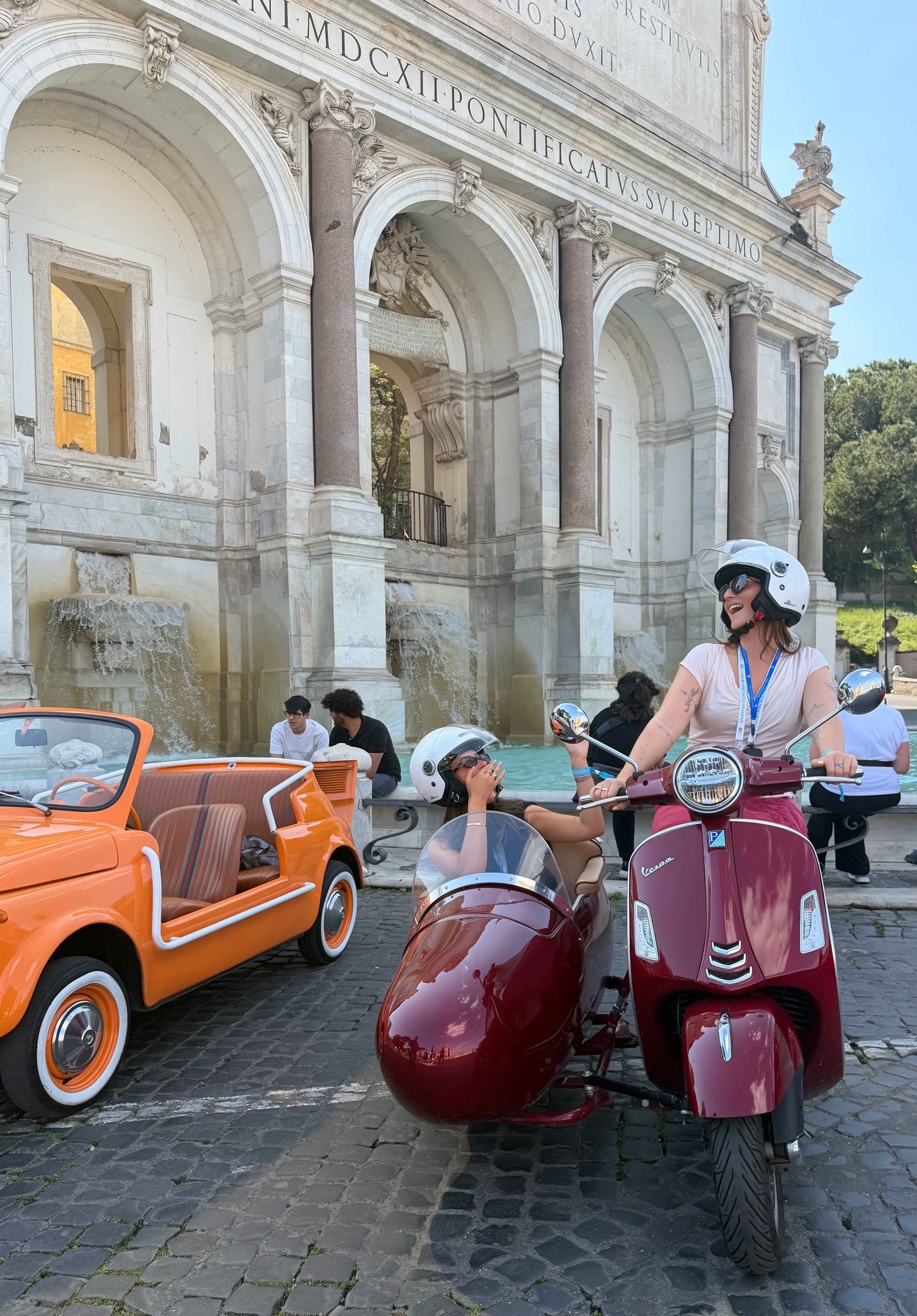 Two women wearing helmets ride a burgundy Vespa with a sidecar beside an orange vintage car in front of a monumental fountain in Rome on a sunny day.