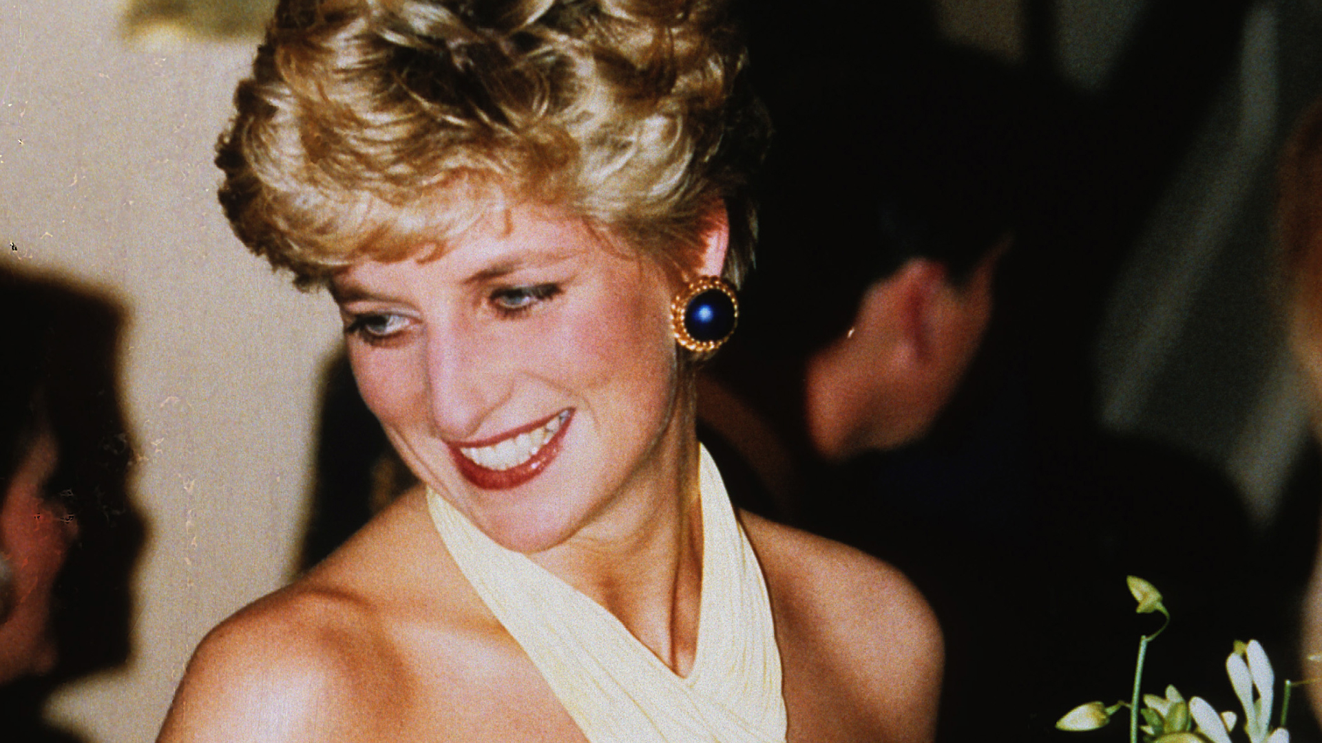 Princess Diana smiles while wearing a pale yellow halter dress and large blue earrings