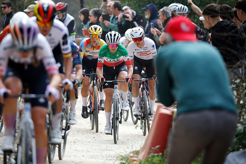 A long shot, taken at Strade Bianche Women 2026, showing blurred riders in the foreground and then focusing in on Elisa Longo Borghini, Lotte Kopecky and Kasia Niewiadoma-Phinney behind