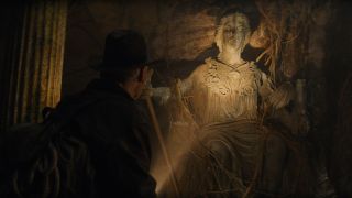 Still from the movie Indiana Jones and the Dial of Destiny. Here we see Indiana Jones shining a torch on an old marble statue of a woman who has snake detailing on her dress. There are lots of plants and branches overgrowing the statue.