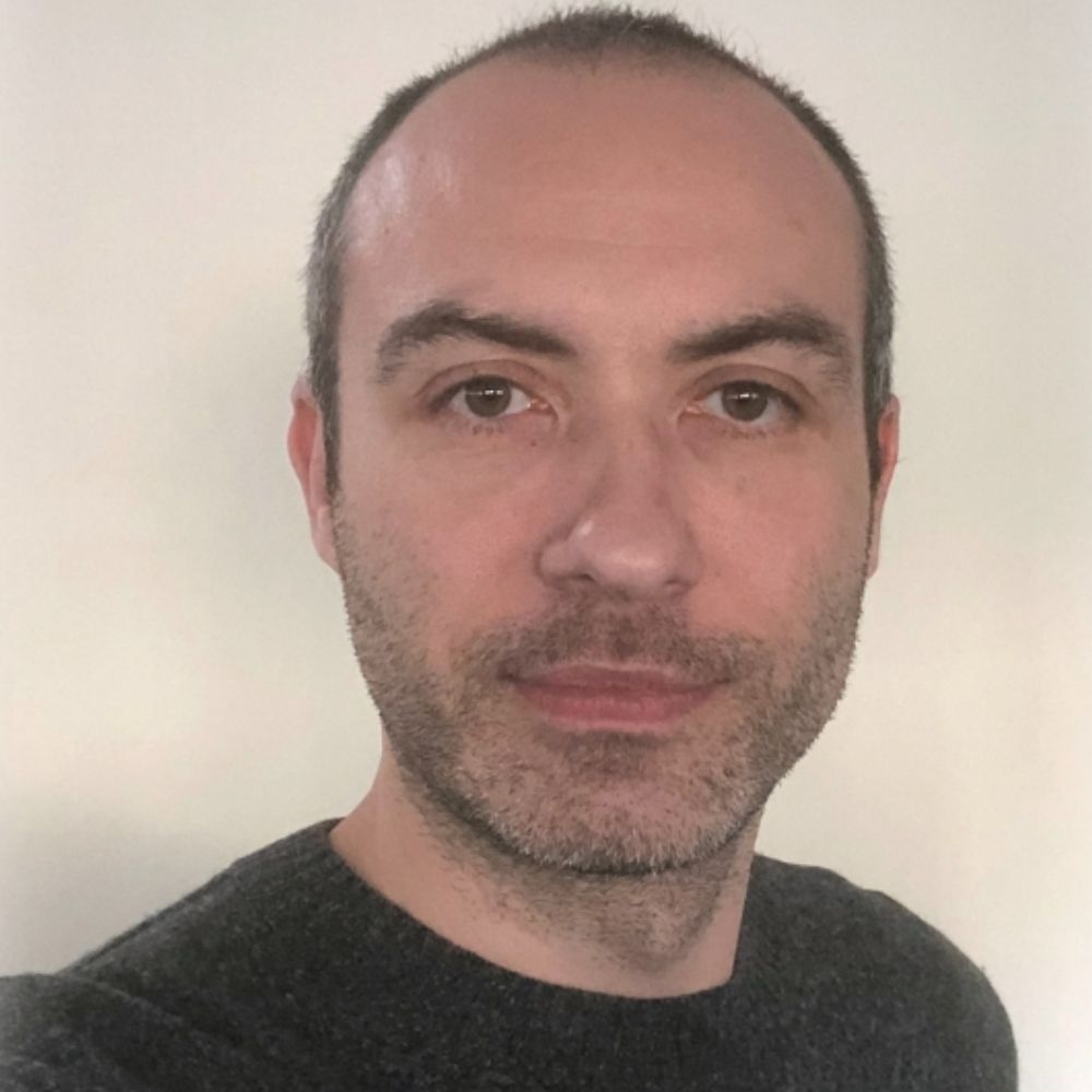 Man with short cropped hair and stubble, wearing a dark grey jumper, close-up headshot against a plain wall.