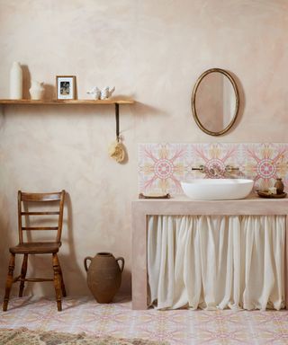 Relaxed bathroom with neutral painted walls, sink with cloth curtain, oval mirror, wooden chair, shelf with decorative ornaments