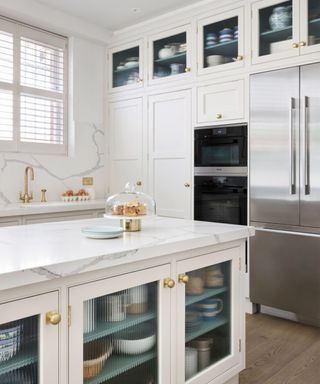 A white kitchen with whitem arble countertops. Tall floor-to-ceiling cabinets along the back wall and gold handle finishes
