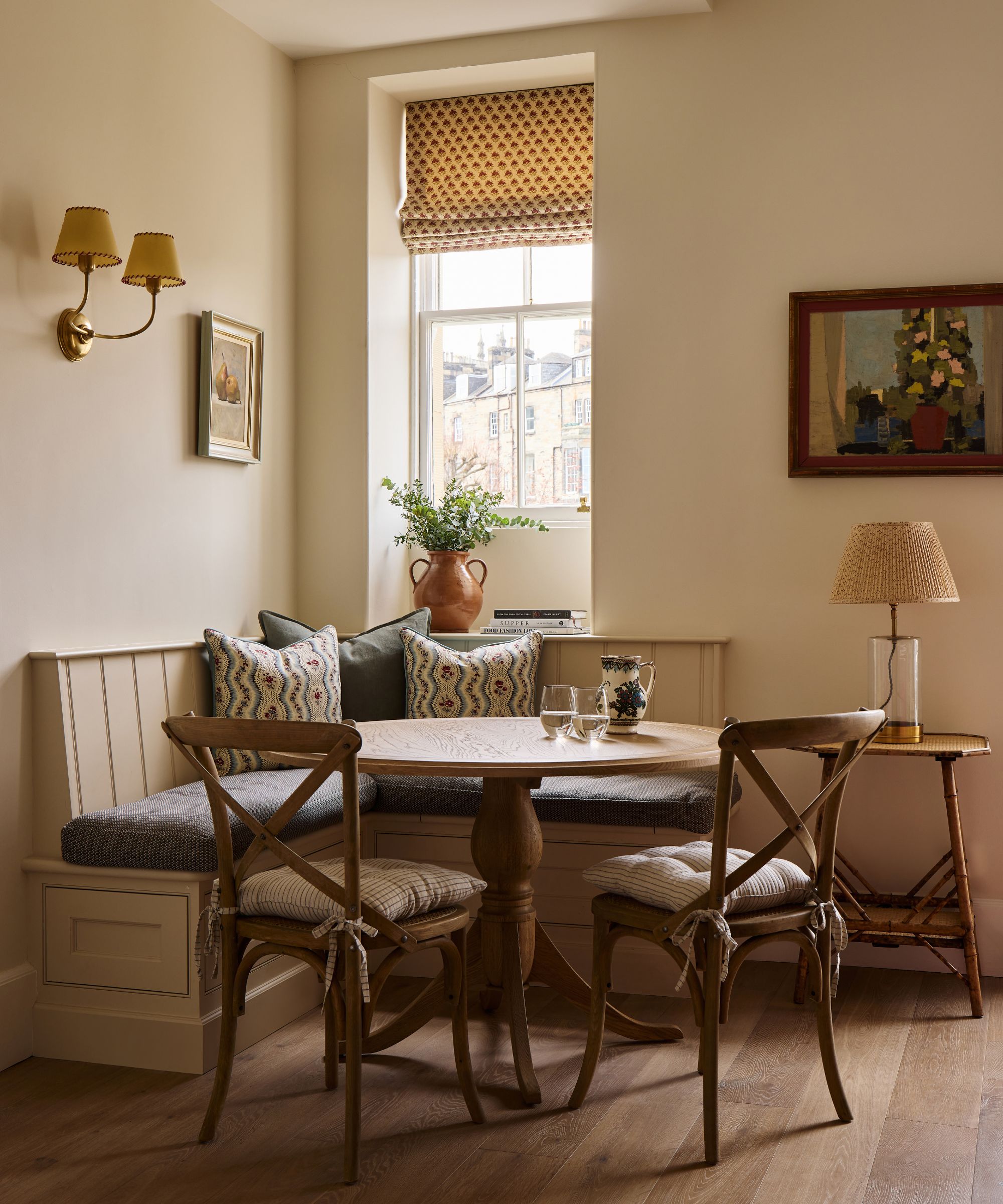 Cozy dining corner with a built-in banquette, patterned throw pillows, a round wooden table, and two wooden cross-back chairs by a window
