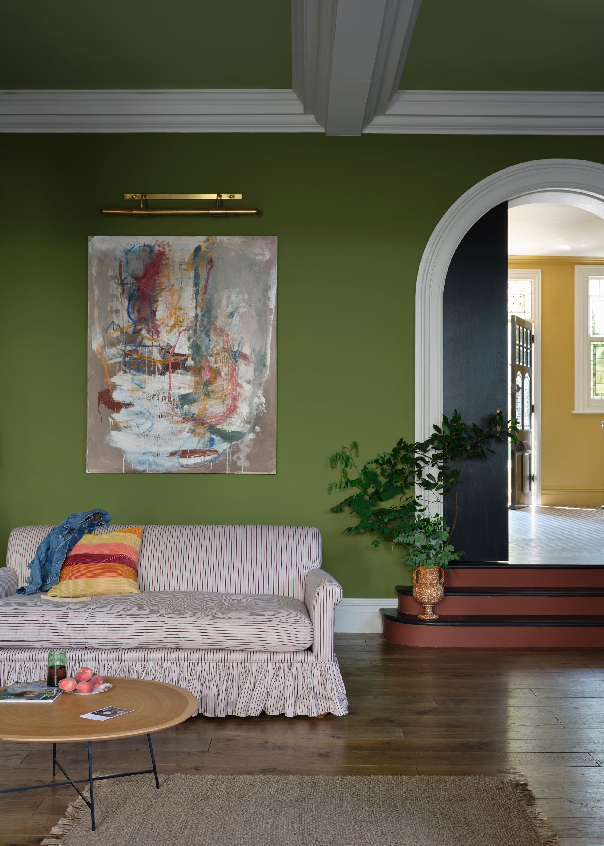 A living room with earthy green walls, white trim, wood floors, and a pale pink and white striped sofa. An arched doorway with stairs leading into a yellow room.