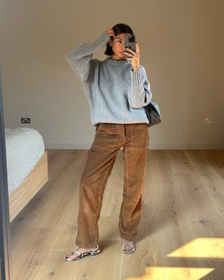 A woman wears a gray chunky sweater, brown corduroy pants, snakeskin sandals.