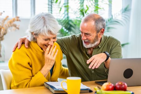 Senior woman looking at household finances notes and expressing surprise and happiness while her husband embracing for the back sitting at home