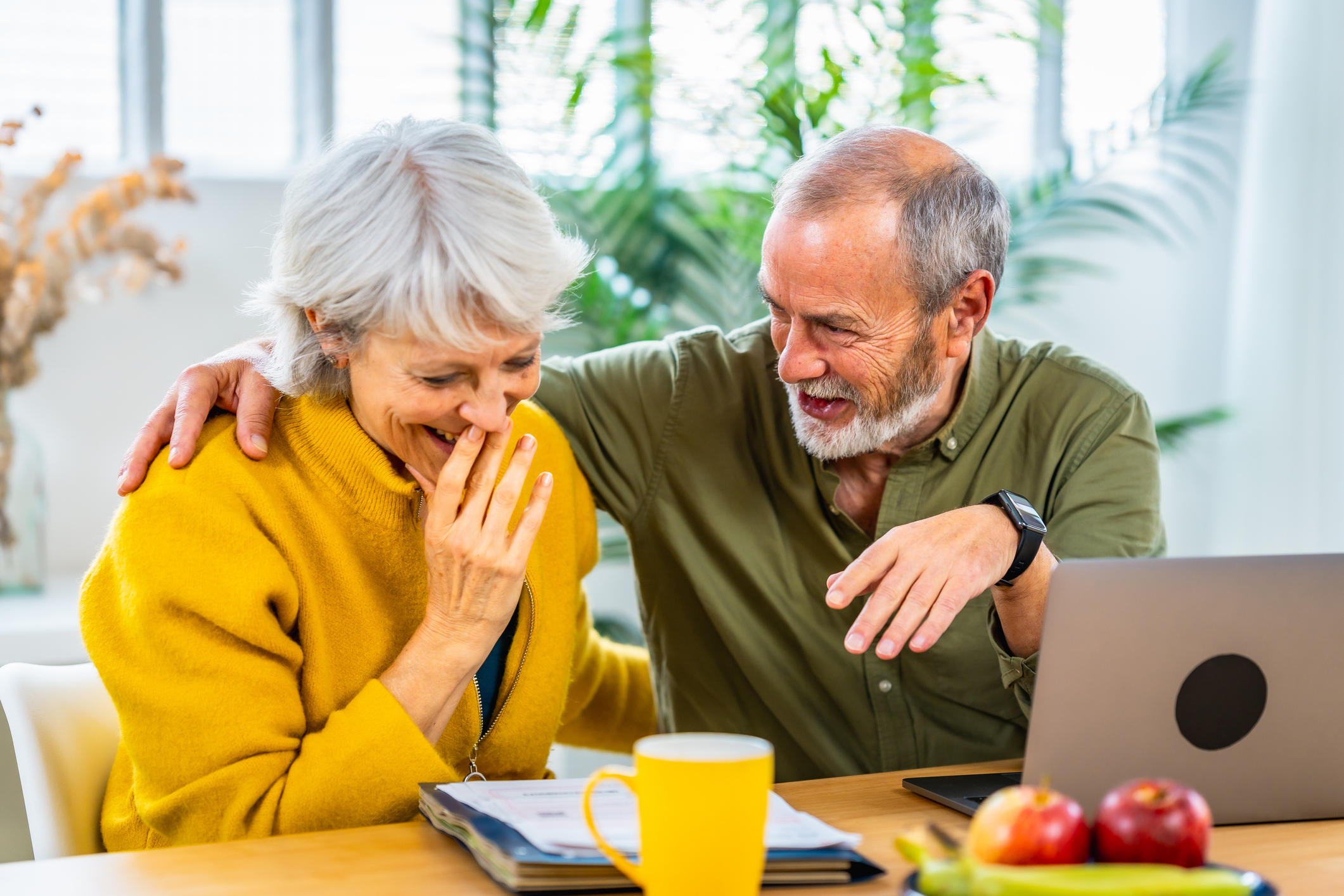 Senior woman looking at household finances notes and expressing surprise and happiness while her husband embracing for the back sitting at home