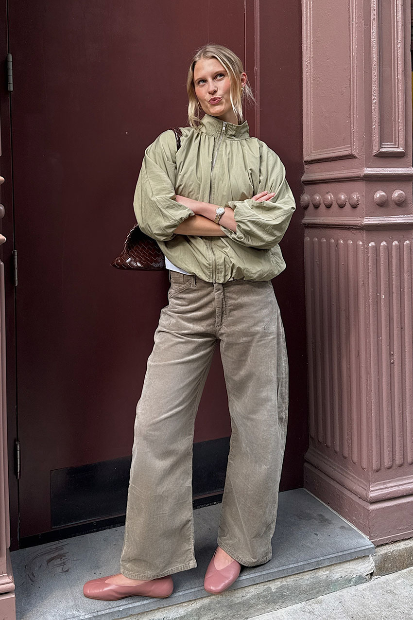 A woman wearing a green funnel-neck jacket, tan barrel jeans, and pink flats.