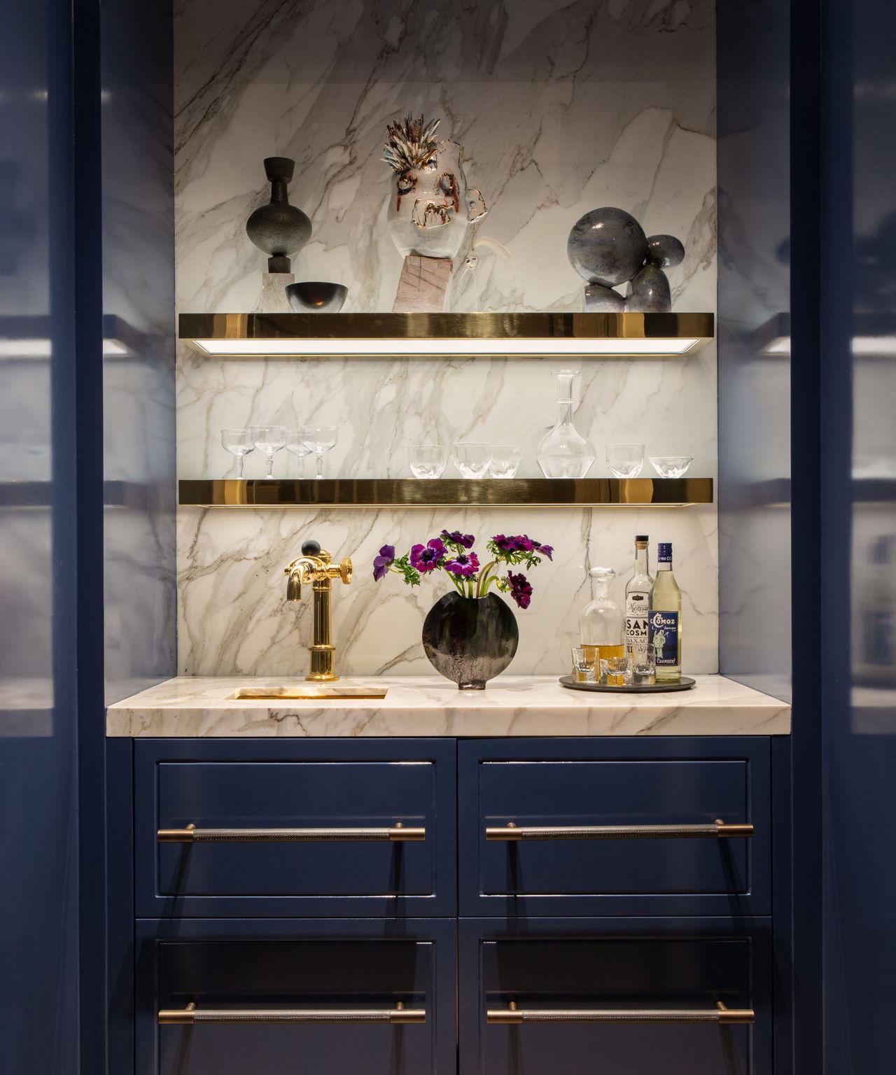 A navy blue home bar with gold shelves and marble backsplash