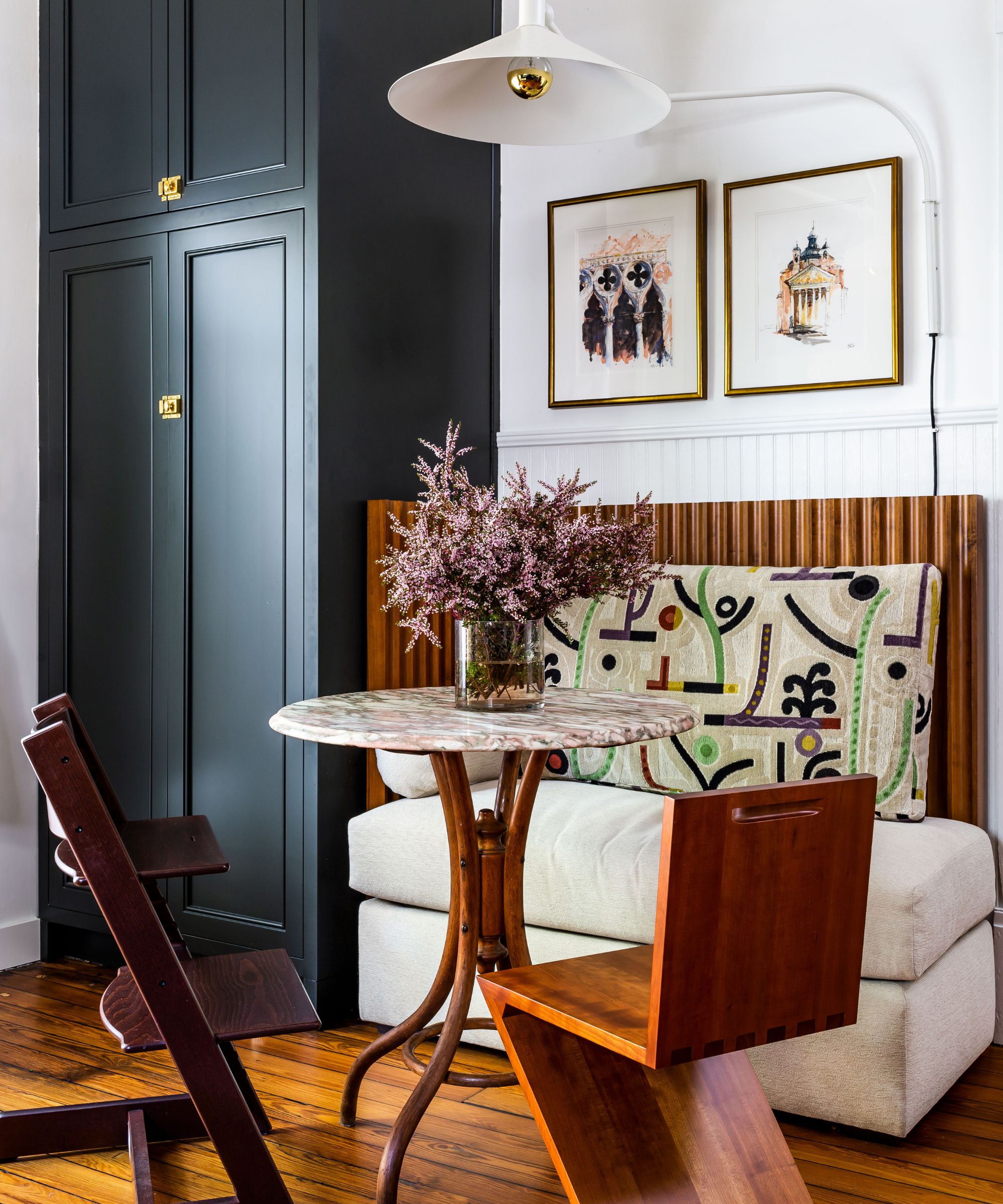 A breakfast nook featuring a marble-topped pedestal table, a modern Z-shaped wood chair, and a cream banquette with a vibrant, abstract-patterned cushion. A tall matte black cabinet and framed sketches on a white wall complete the corner