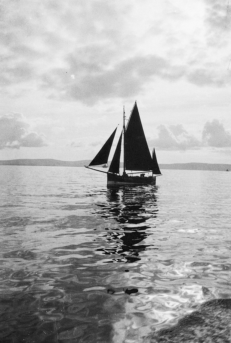 A dark-sailed herring drifter sits becalmed on flat water beneath a wide, cloud-filled sky.