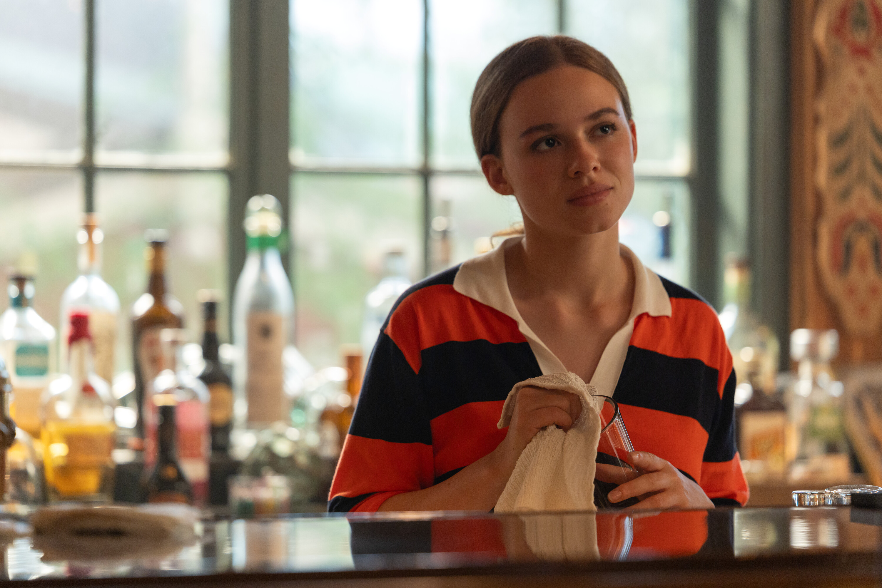 Sadie Soverall as Percy Fraser wearing a striped polo shirt and wiping a glass at a bar in a scene from 'Every Year After.'
