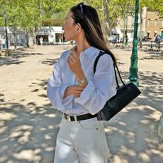 style influencer Felicia Akerstrom poses in a square in St. Tropez wearing black sunglasses on her head, a white button-down shirt, a black Alaia Le Teckel shoulder bag, black belt and ivory jeans