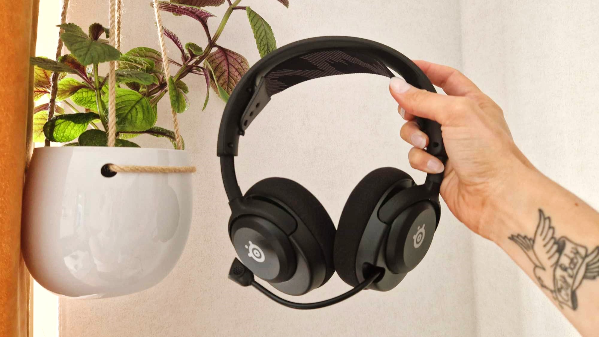 En hand håller upp ett par SteelSeries Arctis Nova 4 framför en hängande krukväxt vid en vit vägg.