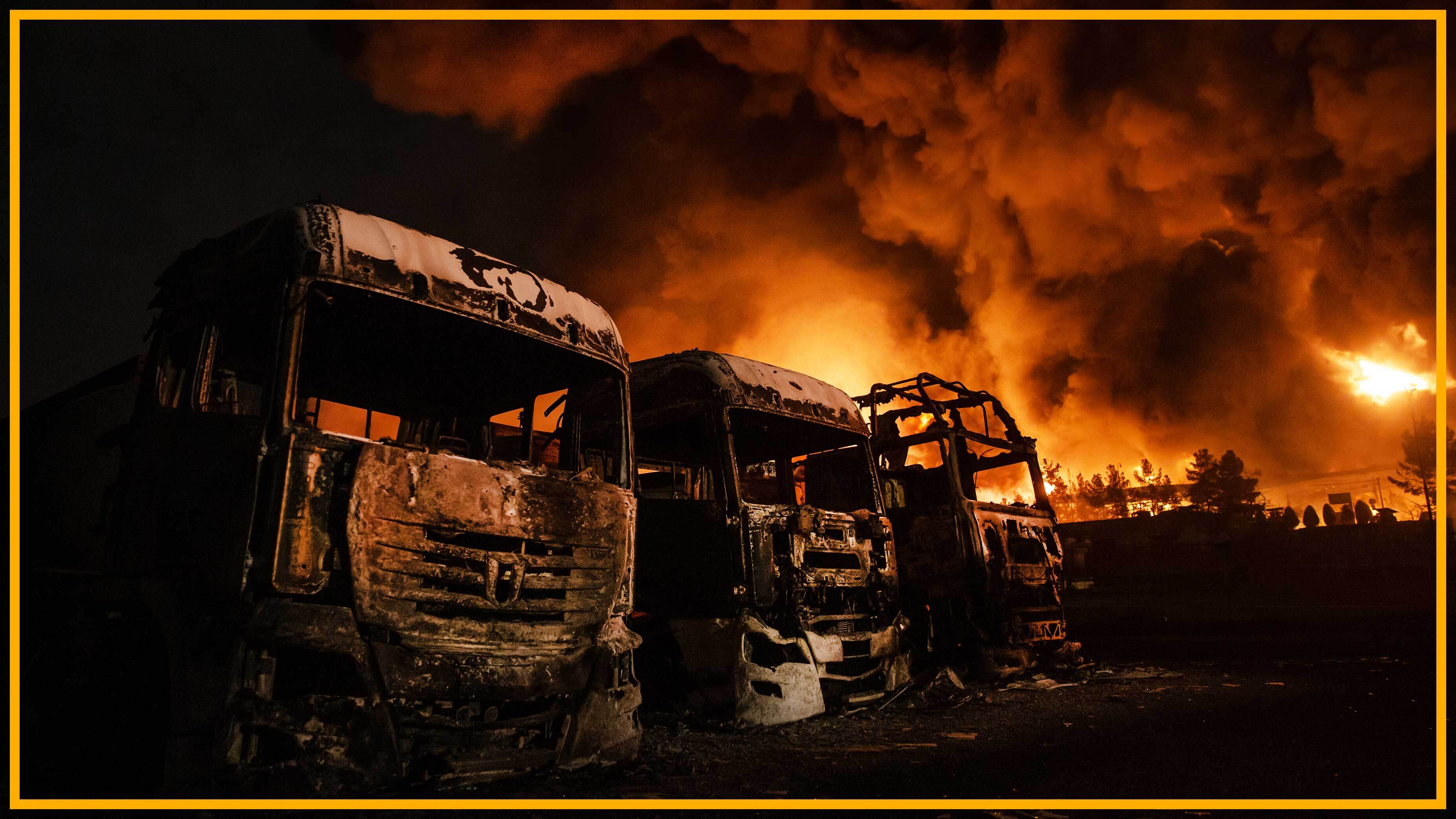 burnt out trucks with fire in the background of an oil depot