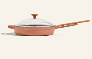 A coral colored cast iron pan with a lid on a plain background.
