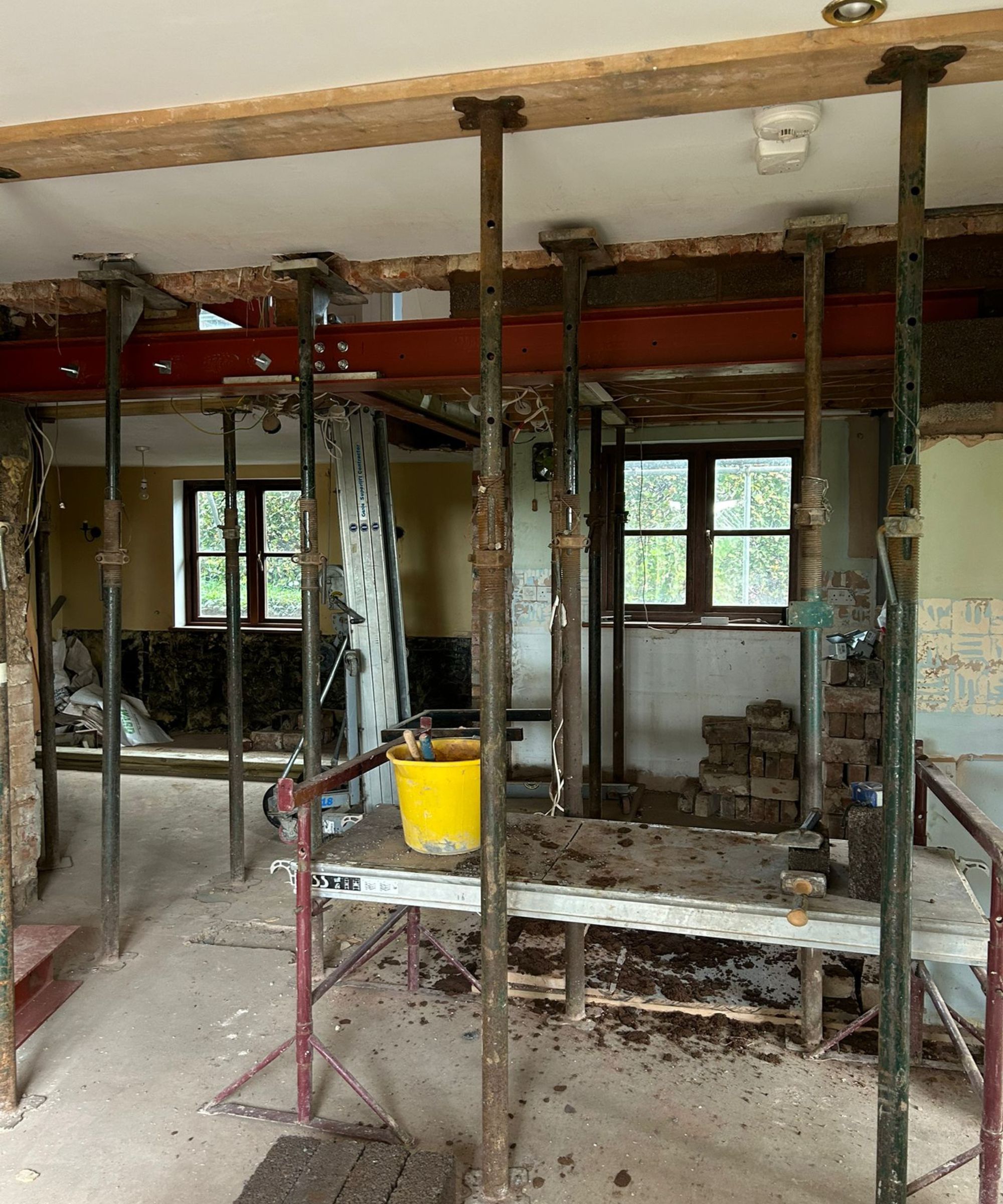 A major structural renovation in progress, with steel beams and rows of temporary acrow props supporting the ceiling. Scaffolding boards, tools and debris fill the space, while stripped walls and exposed wiring show the building mid-transition.