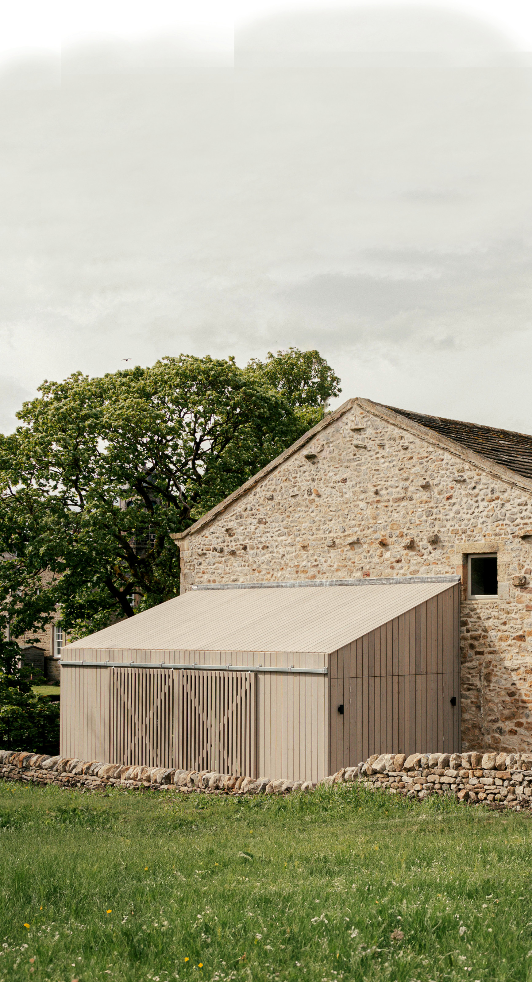 A contemporary wood-clad addition to a barn with pale-hued stone bricks with long grass and trees surrounding it