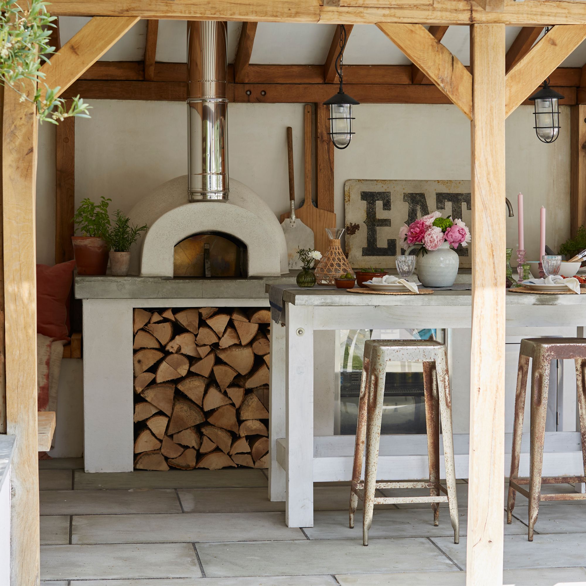 Outdoor kitchen area with cushions beneath wooden roofed pergola with lantern lights and wood stove pizza oven