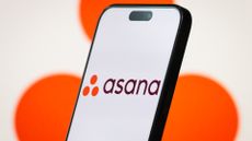 Asana logo and branding pictured on a smartphone screen with company logo blurred in background.
