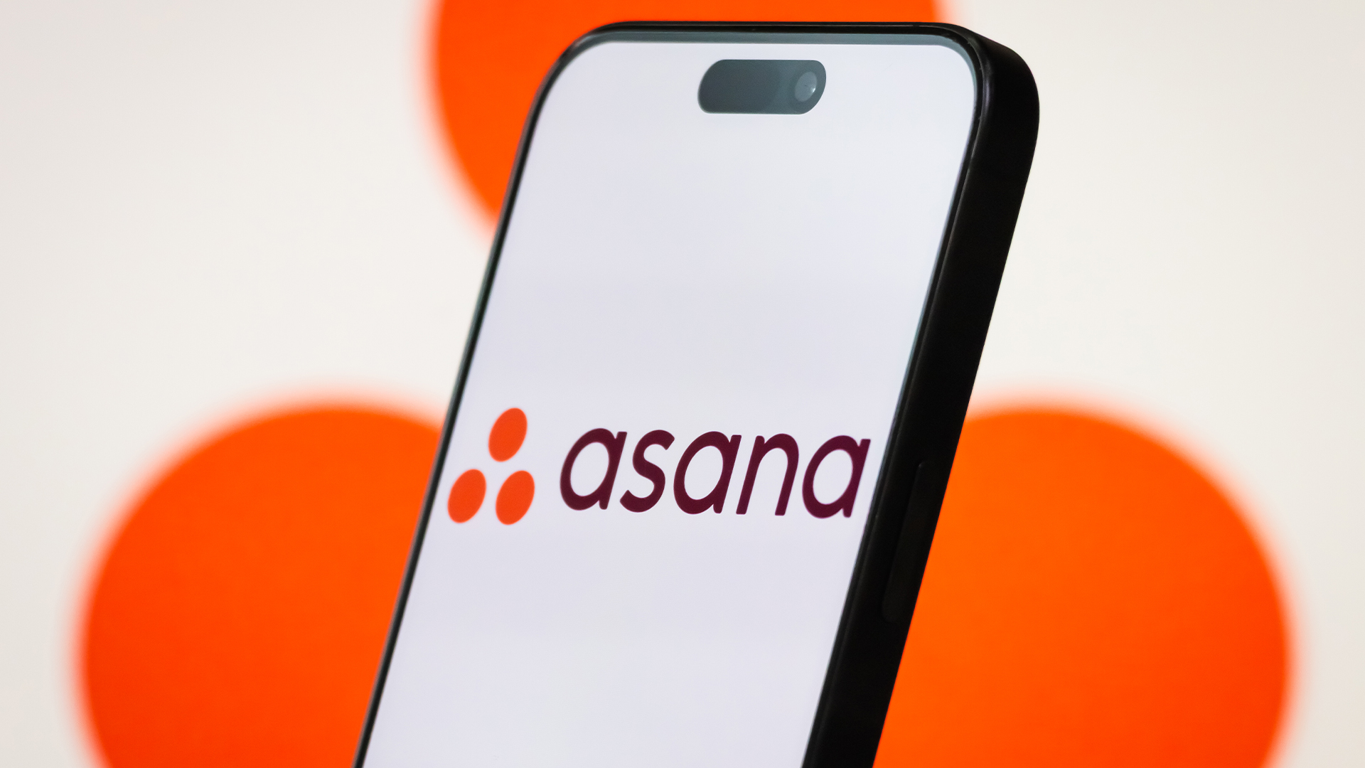 Asana logo and branding pictured on a smartphone screen with company logo blurred in background.