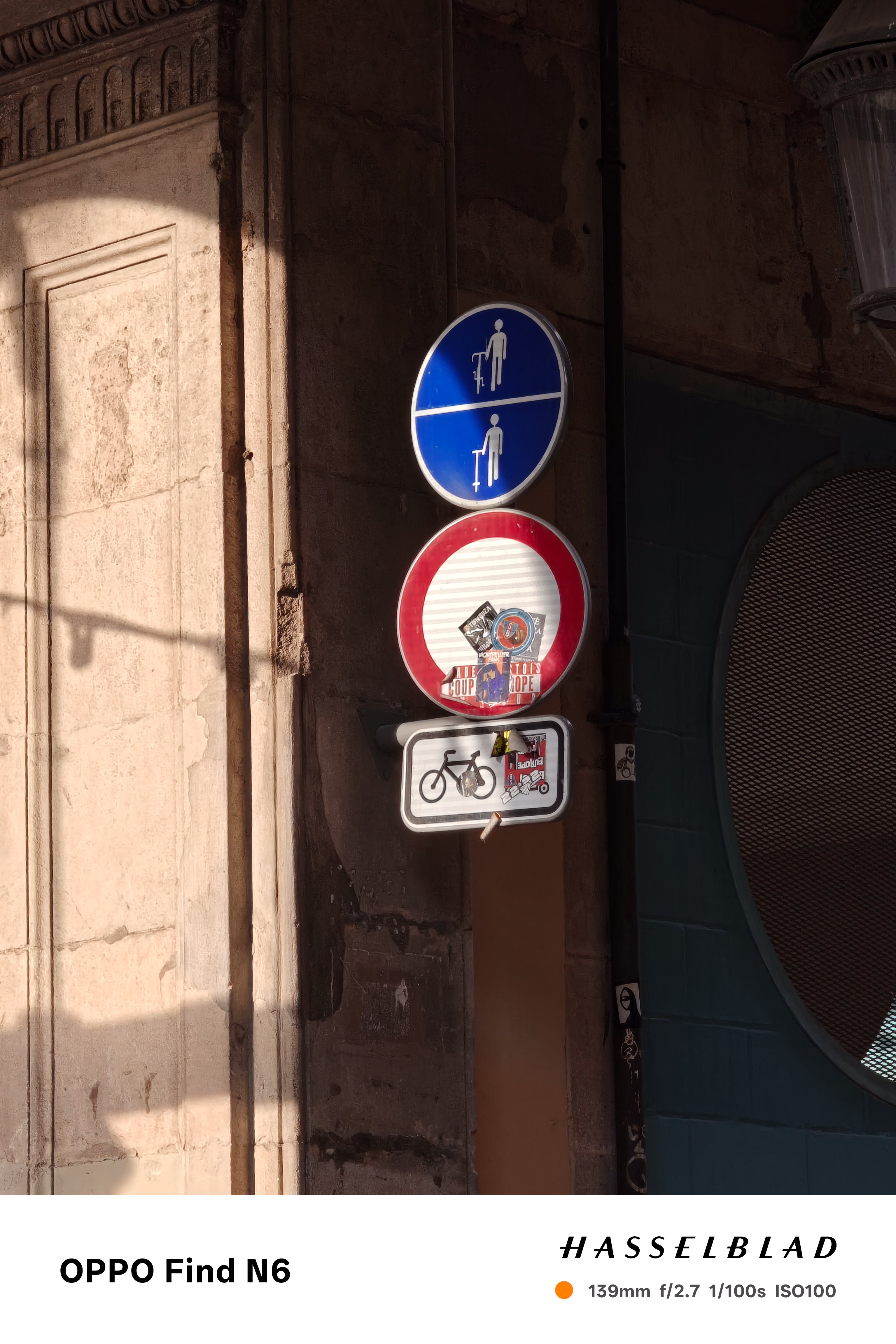 A vertical telephoto shot capturing three stacked European street signs mounted on an old, sun-drenched stone building. The top sign is a blue circle indicating a pedestrian path, the middle is a red and white "no entry" sign covered in stickers, and the bottom is a rectangular white sign with a bicycle icon. Harsh sunlight creates a strong diagonal shadow across the weathered facade.