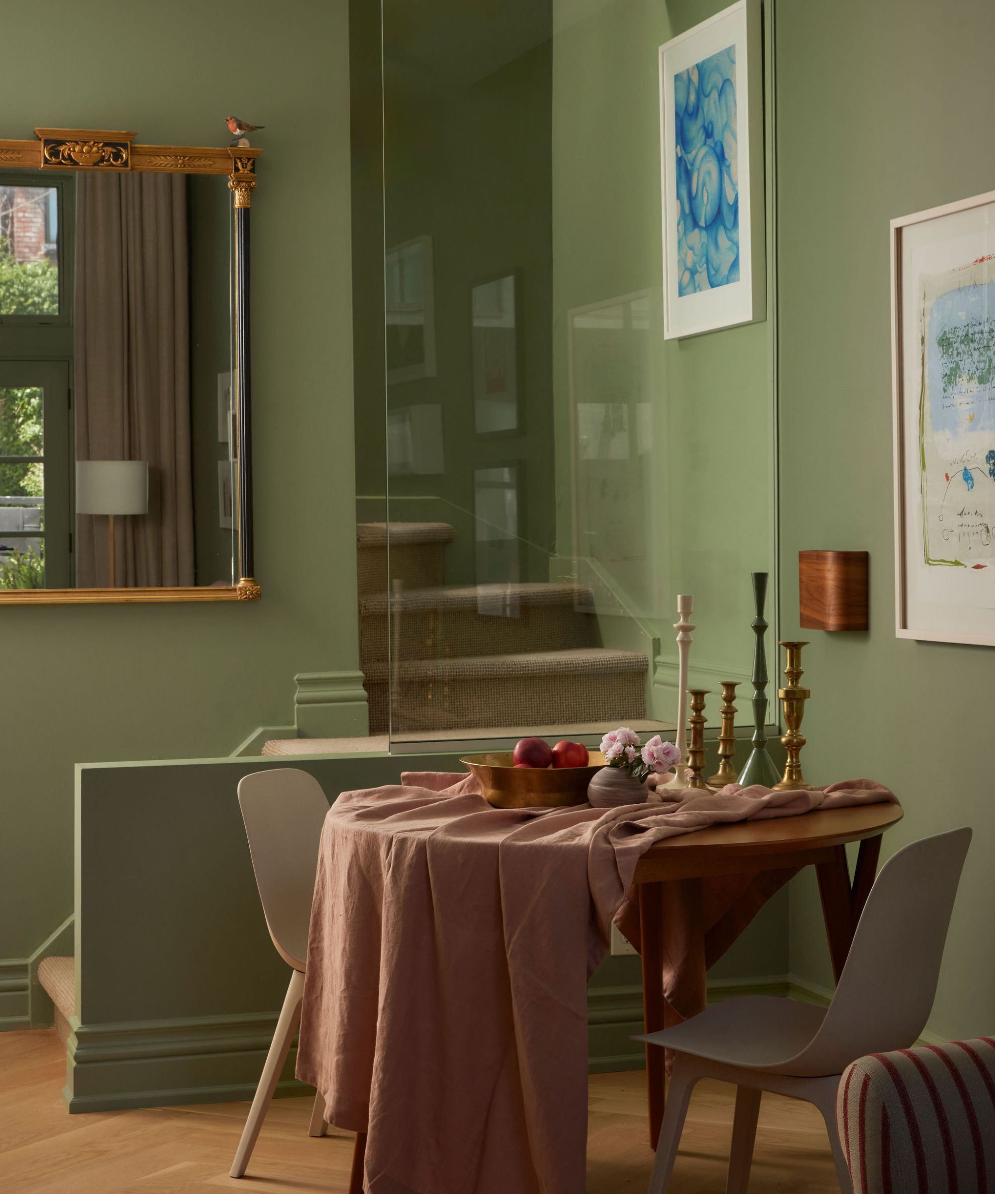 A small dining area with a round wooden table draped in a pink linen cloth, set against a sage green wall and mirrored entryway