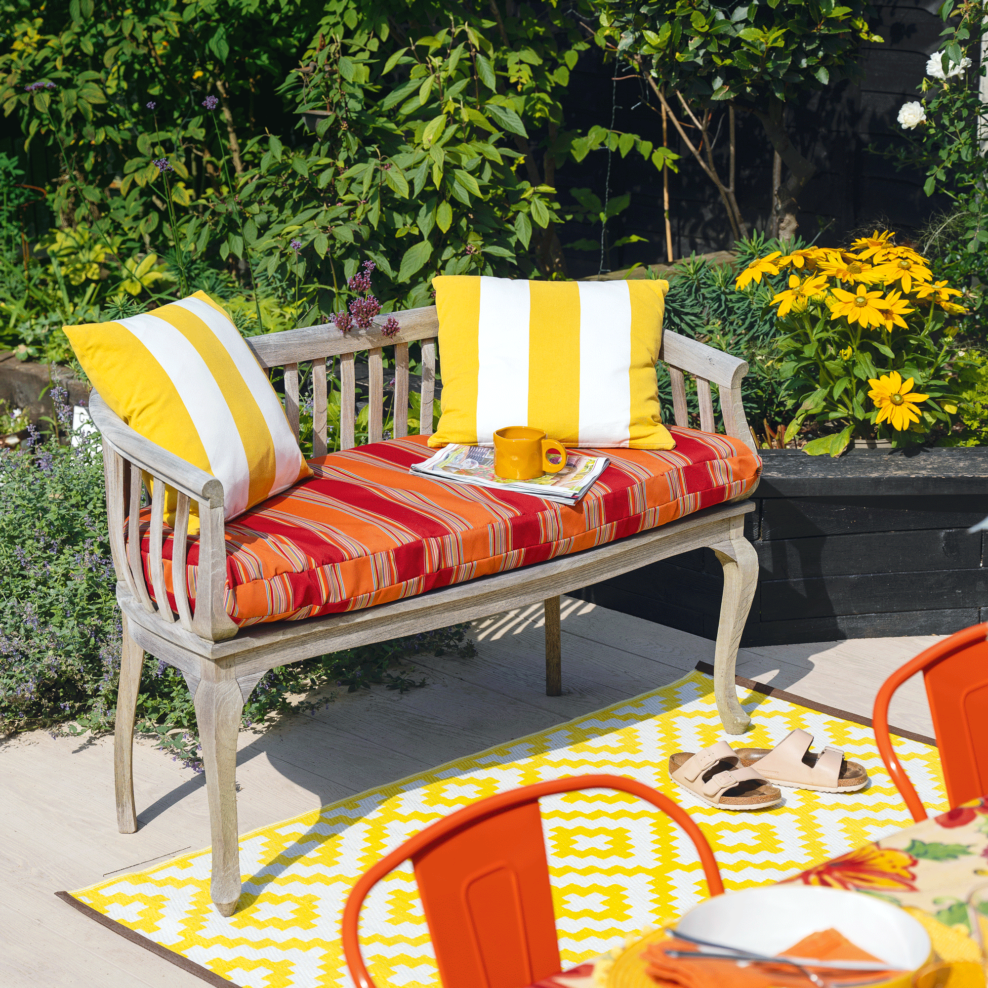 a bench in a garden with an orange and red striped seat pad and yellow and white striped cushions standing on a yellow and white graphic printed outdoor rug