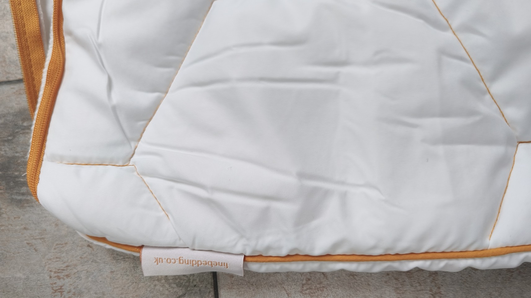 close-up of a white, quilted pillow protector with orange piping and a small fabric tag that reads "finebedding.co.uk."