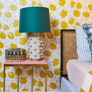 Green lampshade on white bobble lamp base on bedside table in bedroom with yellow printed wallpaper