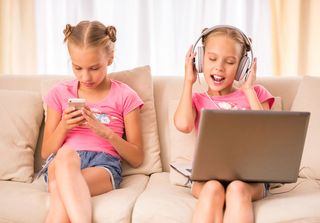 Two girls sitting on the couch using a smartphone and computer.