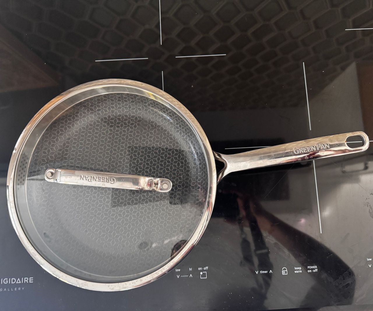 A small stainless steel frying pan with a black interior and a glass lid on a black induction worktop viewed from above.