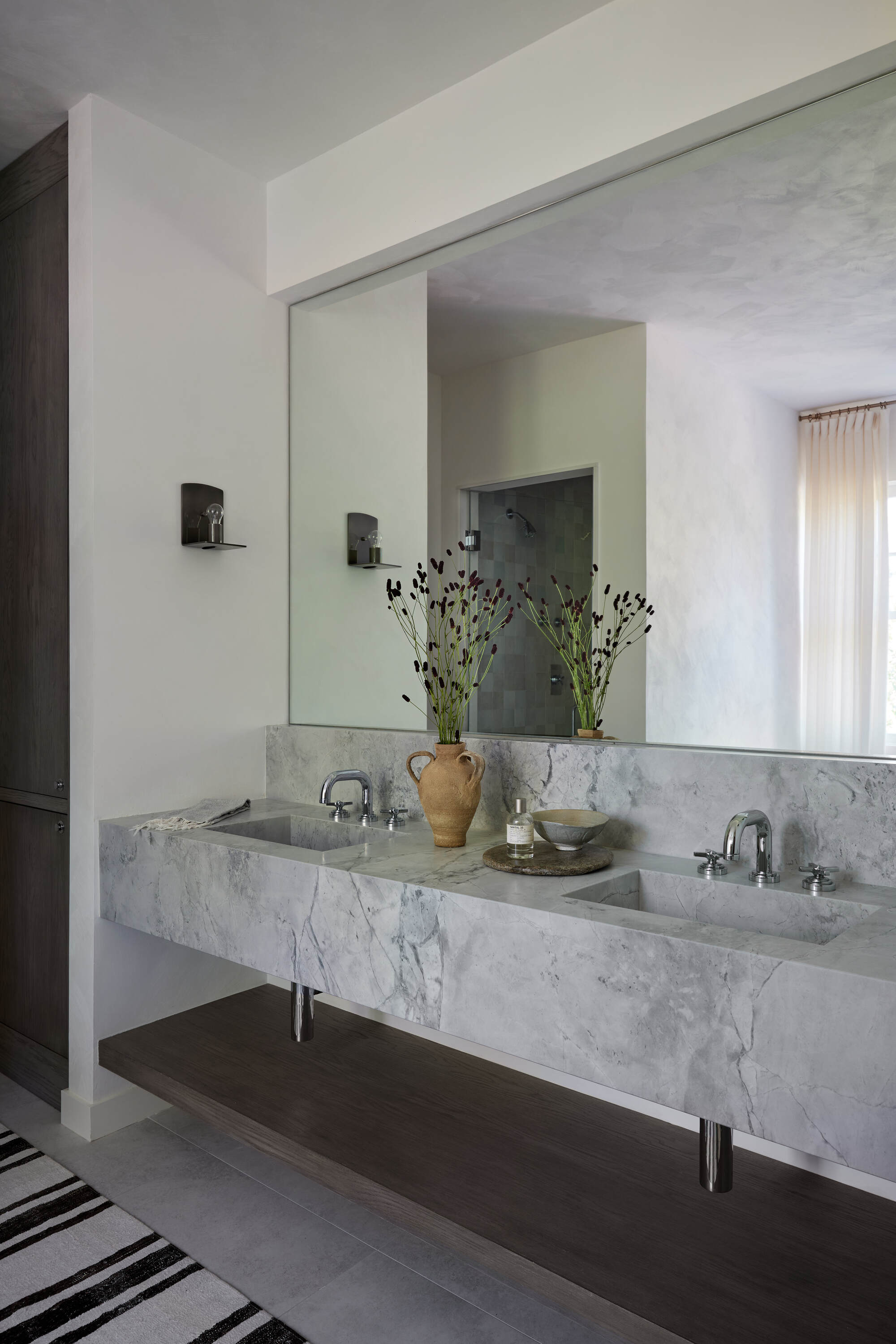 Image of a modern, gray and white bathroom with a large mirror on the wall, double sinks, and a small sconce light.