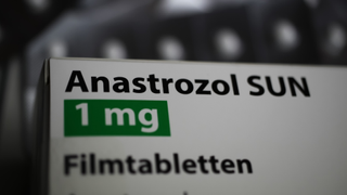 White box containing the drug anastrozol against a blurred, dark background. The drug name is written in black text on the box, as well as "1 mg" written in white text surrounded by a green box.