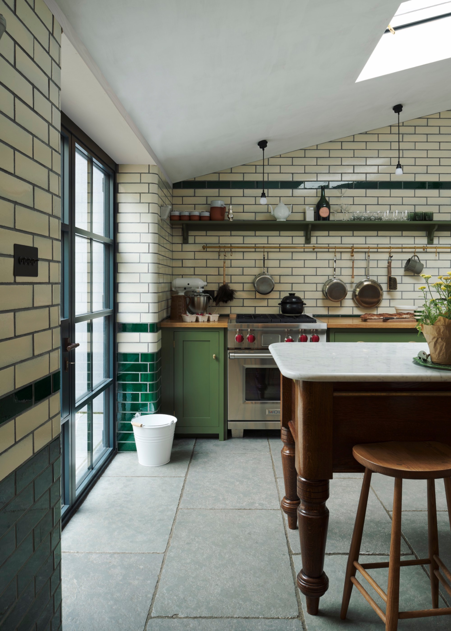 Cream and dark green metro-tiled kitchen with olive cabinetry, stainless steel range, timber island and stone floor.