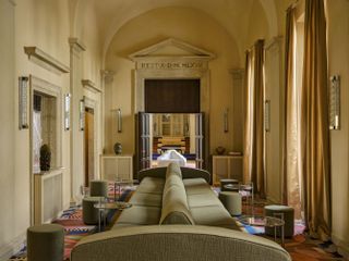 A whimsically furnished hotel lobby features oblong, green upholstered sofas and stools, a colorful patchwork carpet, contemporary glass lighting, and antique architectural volumes.