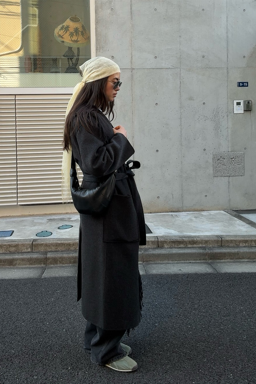 How to wear a bandana is shown in a photo of a woman standing in the street wearing a beige headscarf with a black trench coat, black baggy jeans, olive green sneakers, and a black crossbody bag
