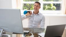 Man sits at a desk, holding a kettlebell on one shoulder. 